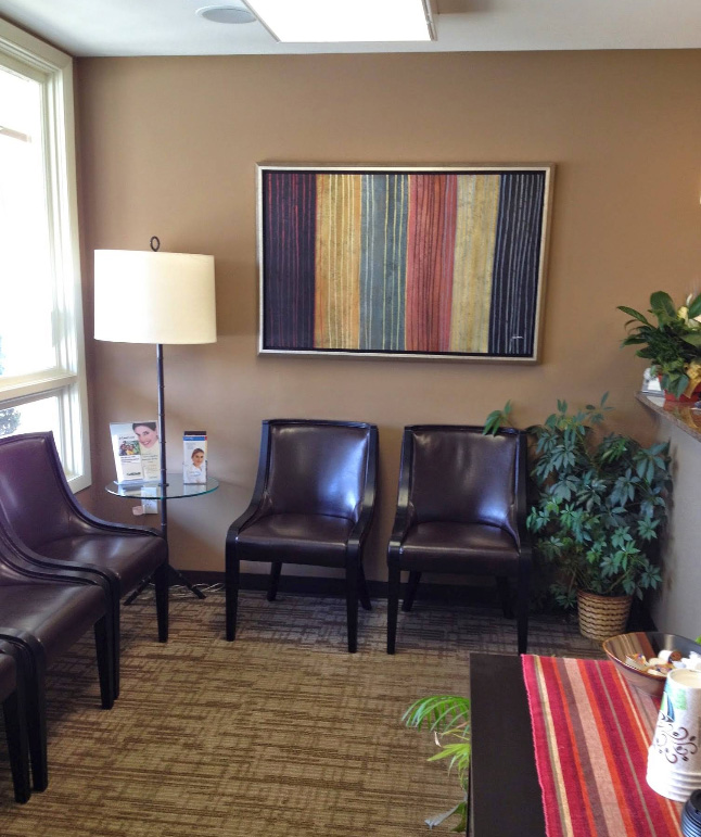 The image shows an interior space, likely a waiting area within a medical office, featuring two chairs, a small table, a framed abstract art piece on the wall, a lamp with a white shade, a potted plant, and a window with blinds partially open, allowing natural light to enter.
