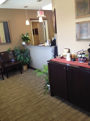 The image shows an interior view of a professional office space with a reception area, featuring a desk, chair, framed artwork on the wall, potted plants, and a well-maintained appearance.
