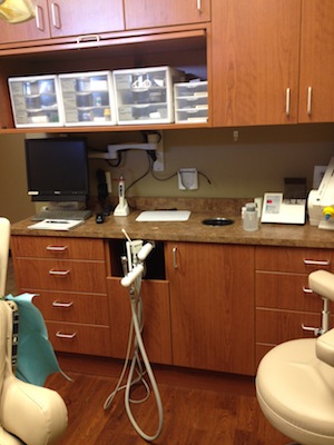 The image shows an interior view of a dental office with a clean and organized appearance, featuring a reception desk with a computer monitor, dental equipment on shelves, and a large window allowing natural light into the room.