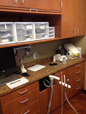 The image shows a clean, well-organized dental office with a countertop containing various dental equipment and supplies, and a computer monitor displaying what appears to be patient records or treatment plans.