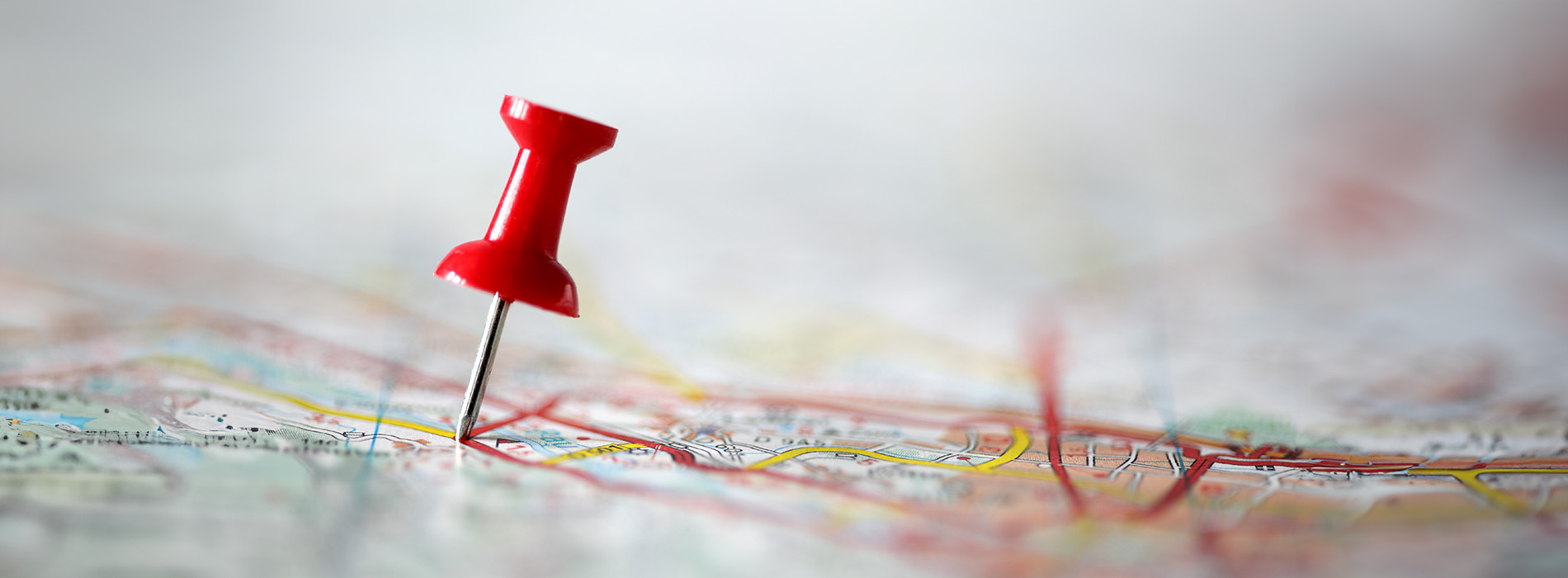 The image features a close-up of a map with a red pushpin inserted into it, which is placed on a blurred background that appears to be indoors with a focus on the pin and the map.