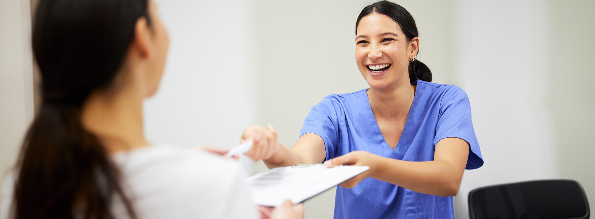 The image shows a woman in scrubs smiling at a person she s handing something to, with another individual looking on from behind.